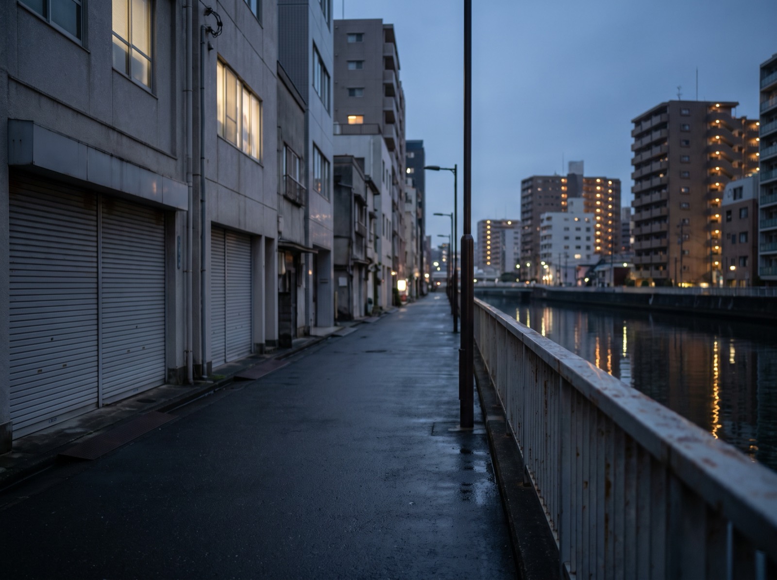 A narrow riverside street at dusk, a plain railing and dark water along one side.