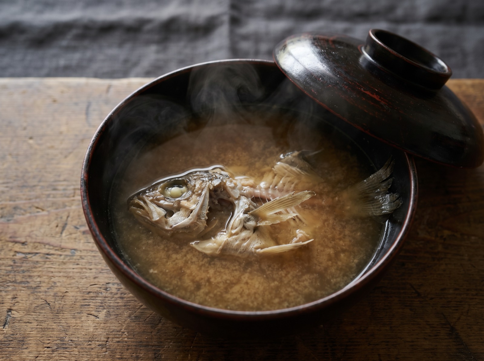 Misoshiru, a soup bowl with its lid just lifted to reveal cloudy broth and fish bones.