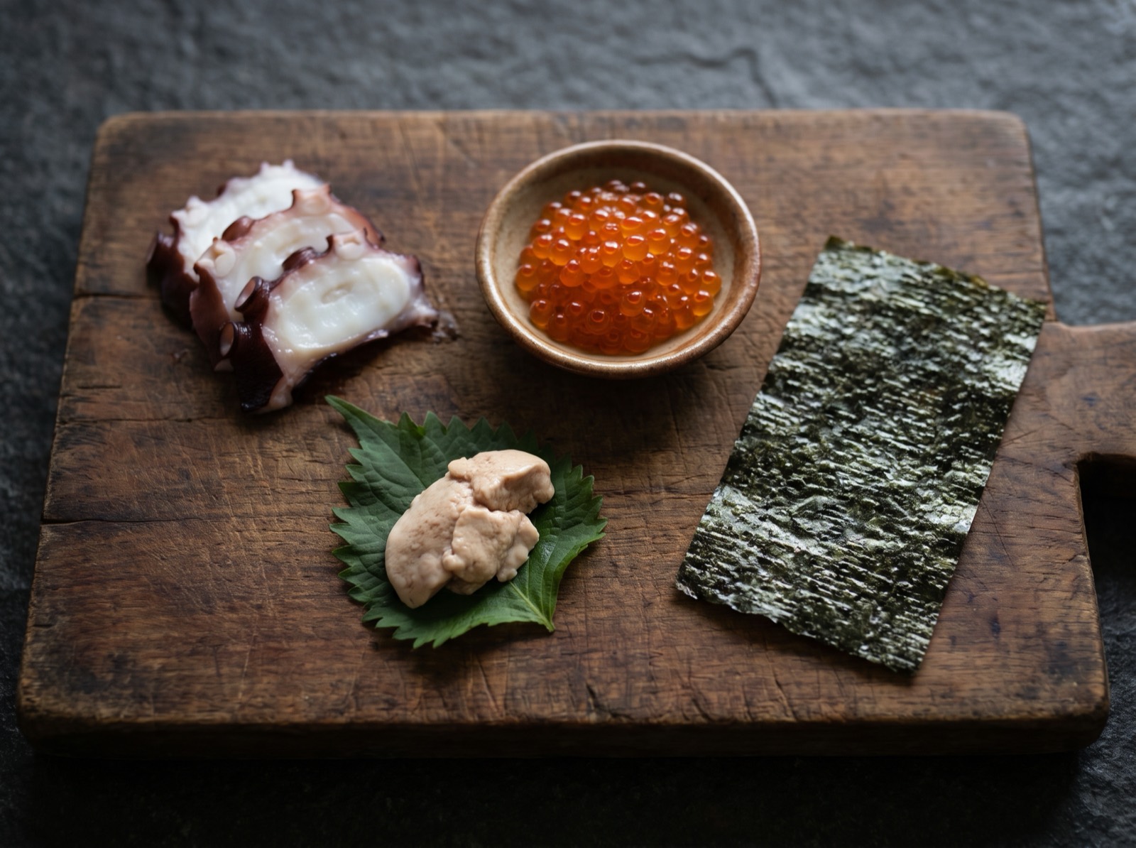 Sliced boiled octopus, salmon roe, a filefish liver and a sheet of toasted nori on a dark wooden board.