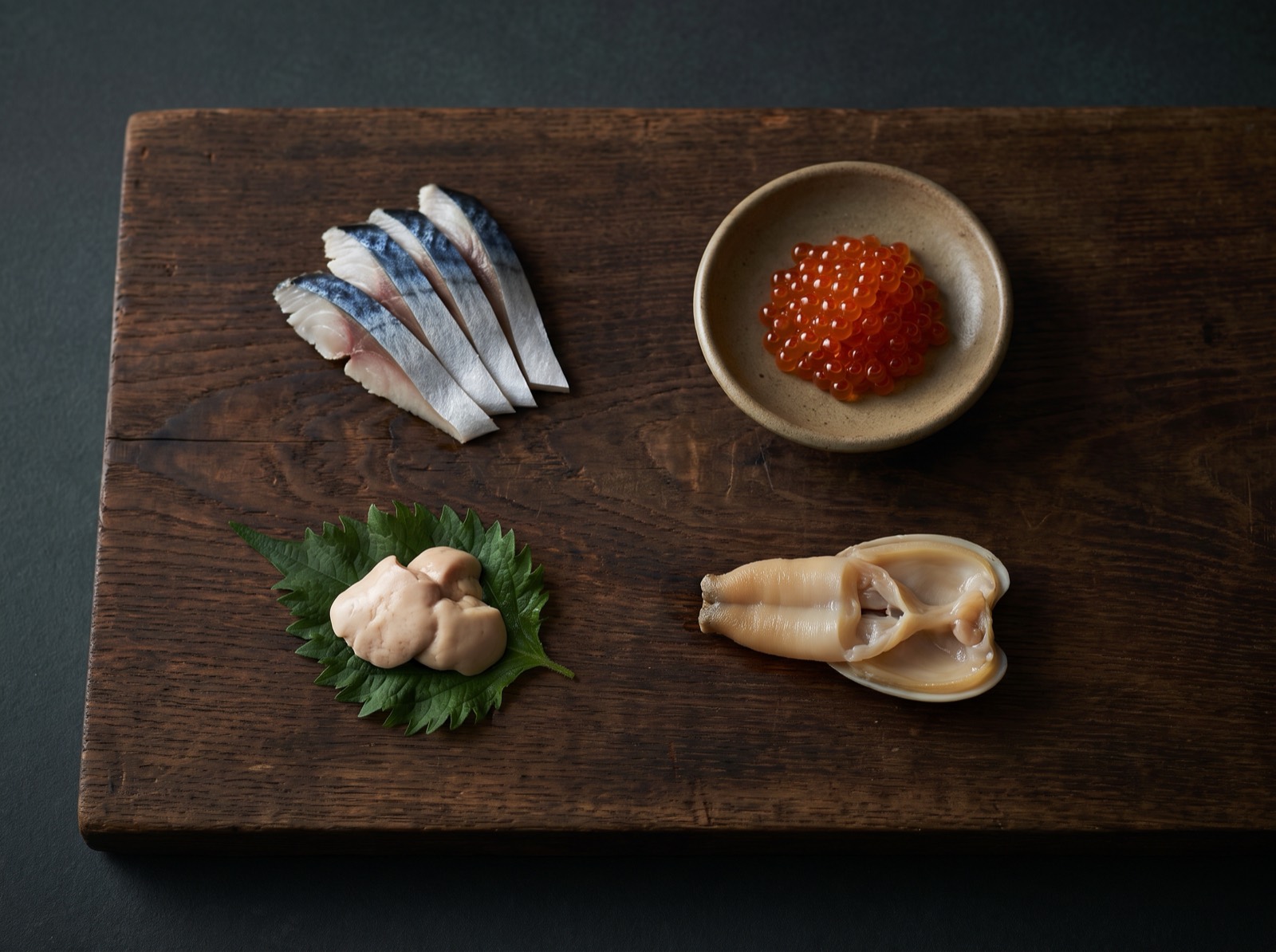 Sliced saury, salmon roe, a filefish liver and a clam siphon on a dark wooden board.