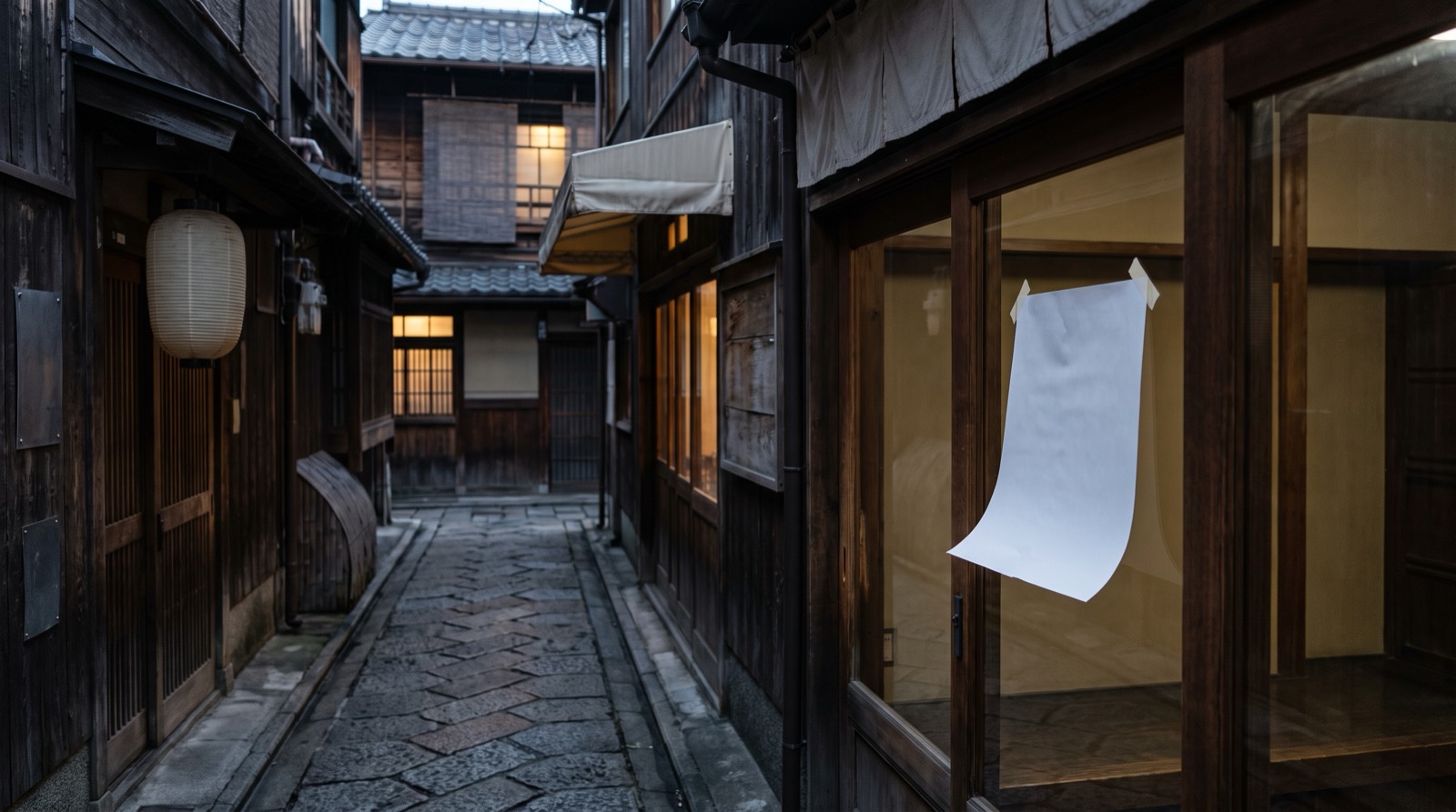 A narrow old shopping street at dusk, a sheet of paper taped inside a glass door.