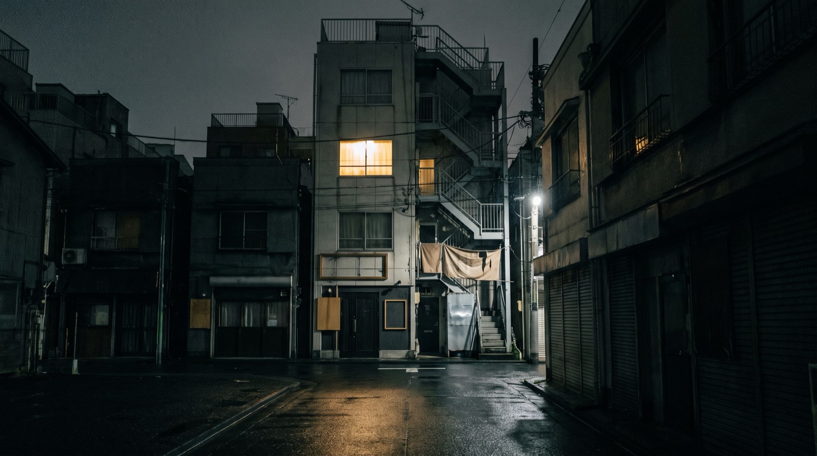 A narrow multi-tenant building at night, one lit window on an upper floor.